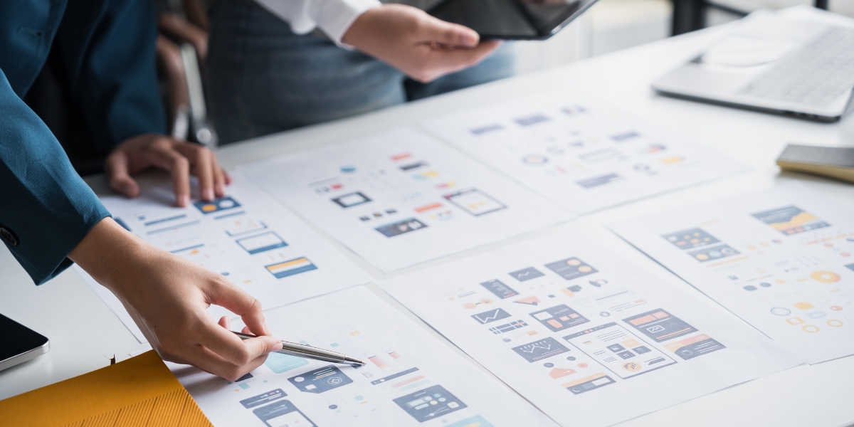 A designer and team members review wireframe mockups and interface layouts spread across a table, planning exceptional user experience across all devices with collaborative sketches and prototypes.